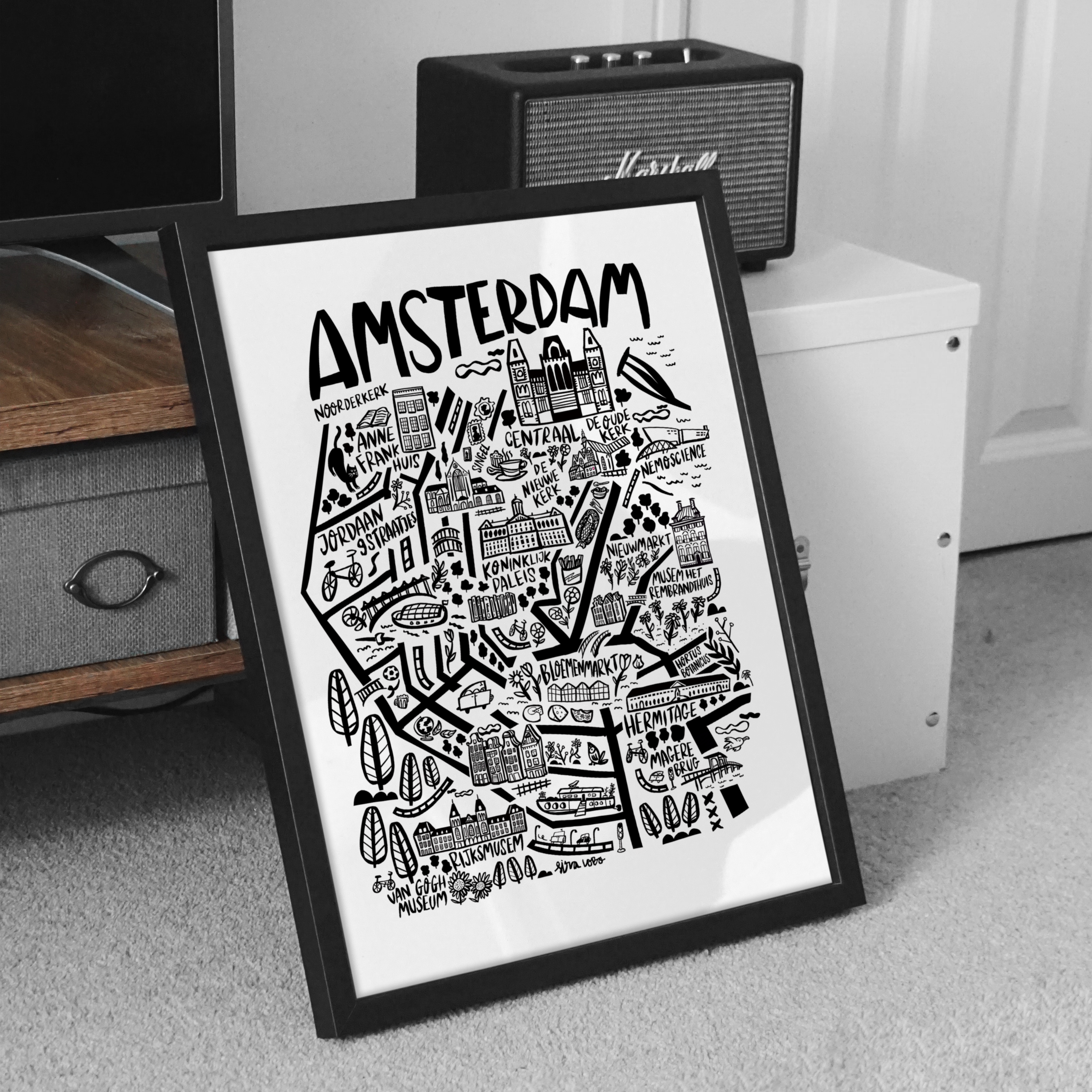 Framed map of Amsterdam on a white wall with a retro radio and wooden shelf in the background.