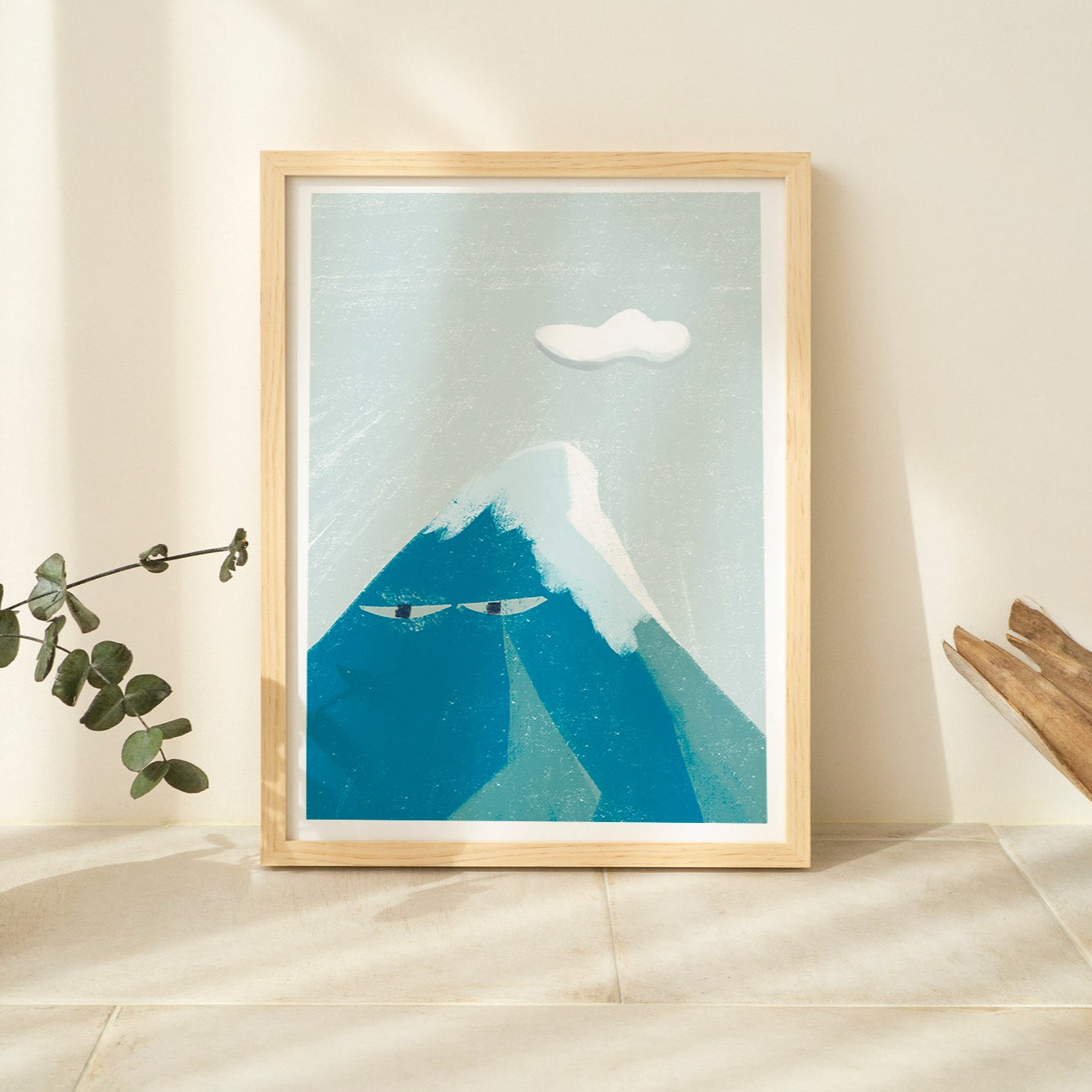 Framed artwork of a mountain with a plant and wooden object on a light-colored floor.