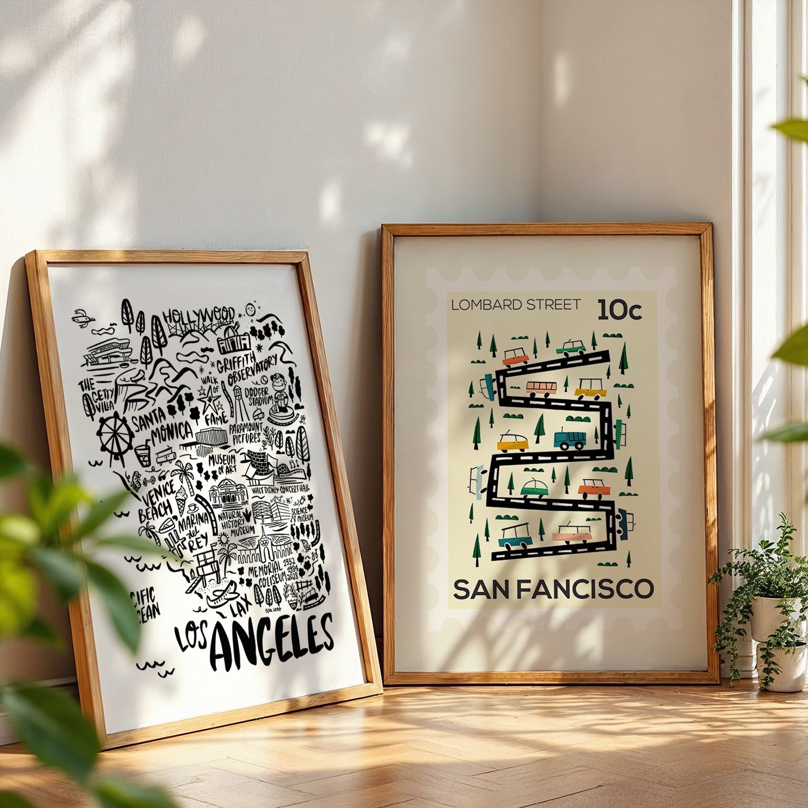 Two framed maps of Los Angeles and San Francisco on a wooden floor with plants around.