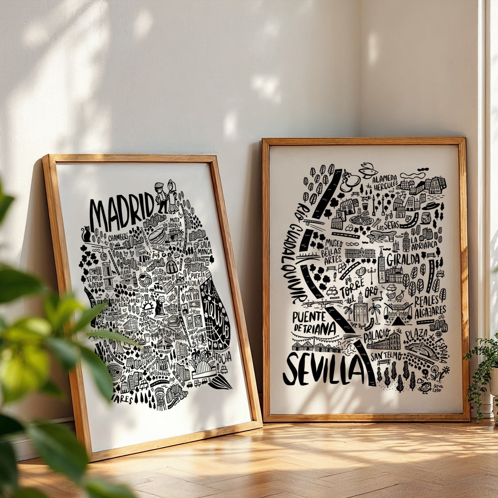 Two framed black and white maps of Madrid and Sevilla on a light wooden floor with plants around.