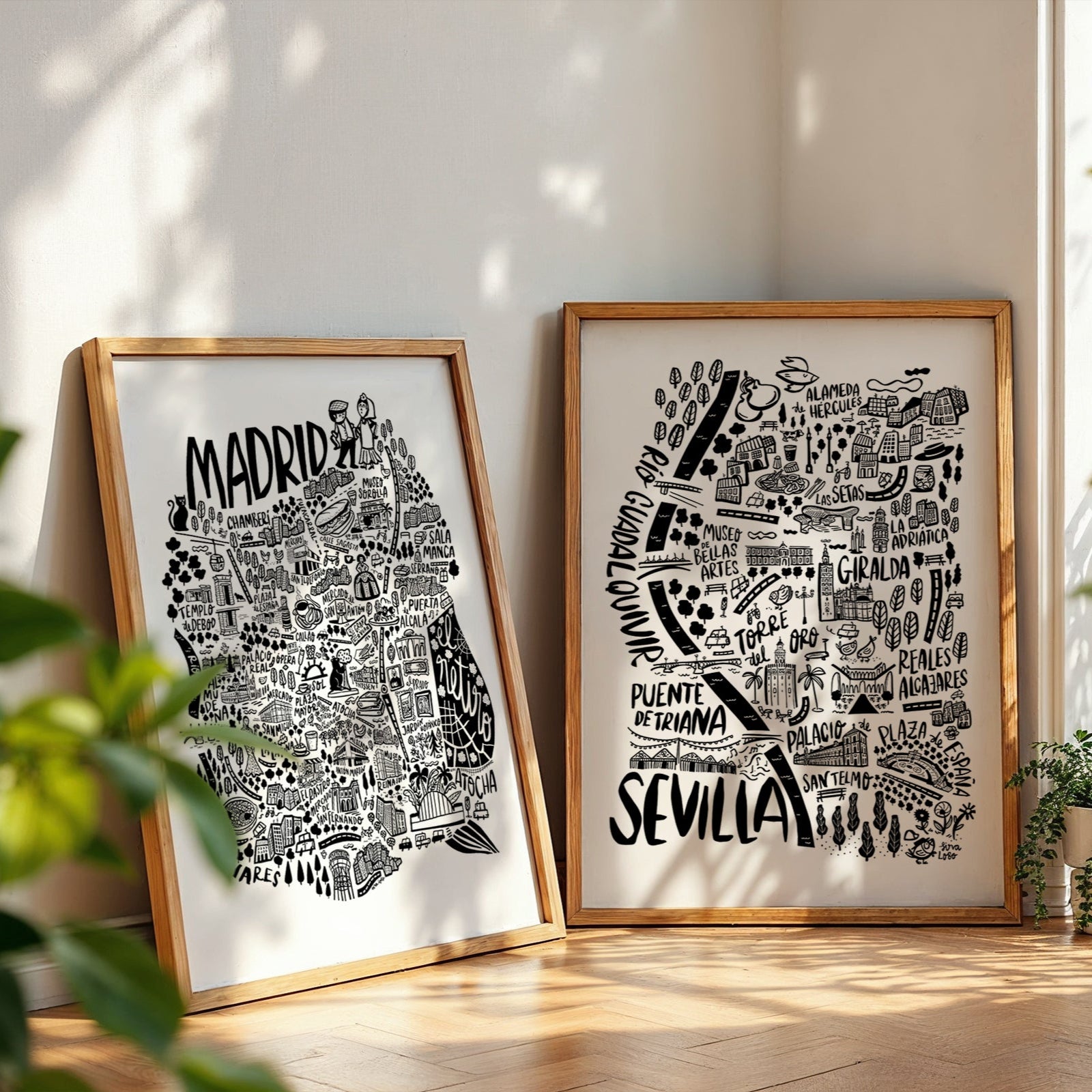 Two framed prints of city names, 'Madrid' and 'Sevilla', on a wall with plants and sunlight.