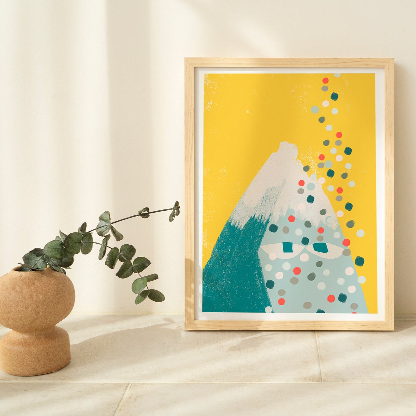 Framed abstract art print with mountain and polka dot design on a yellow background, placed on a light wooden shelf.