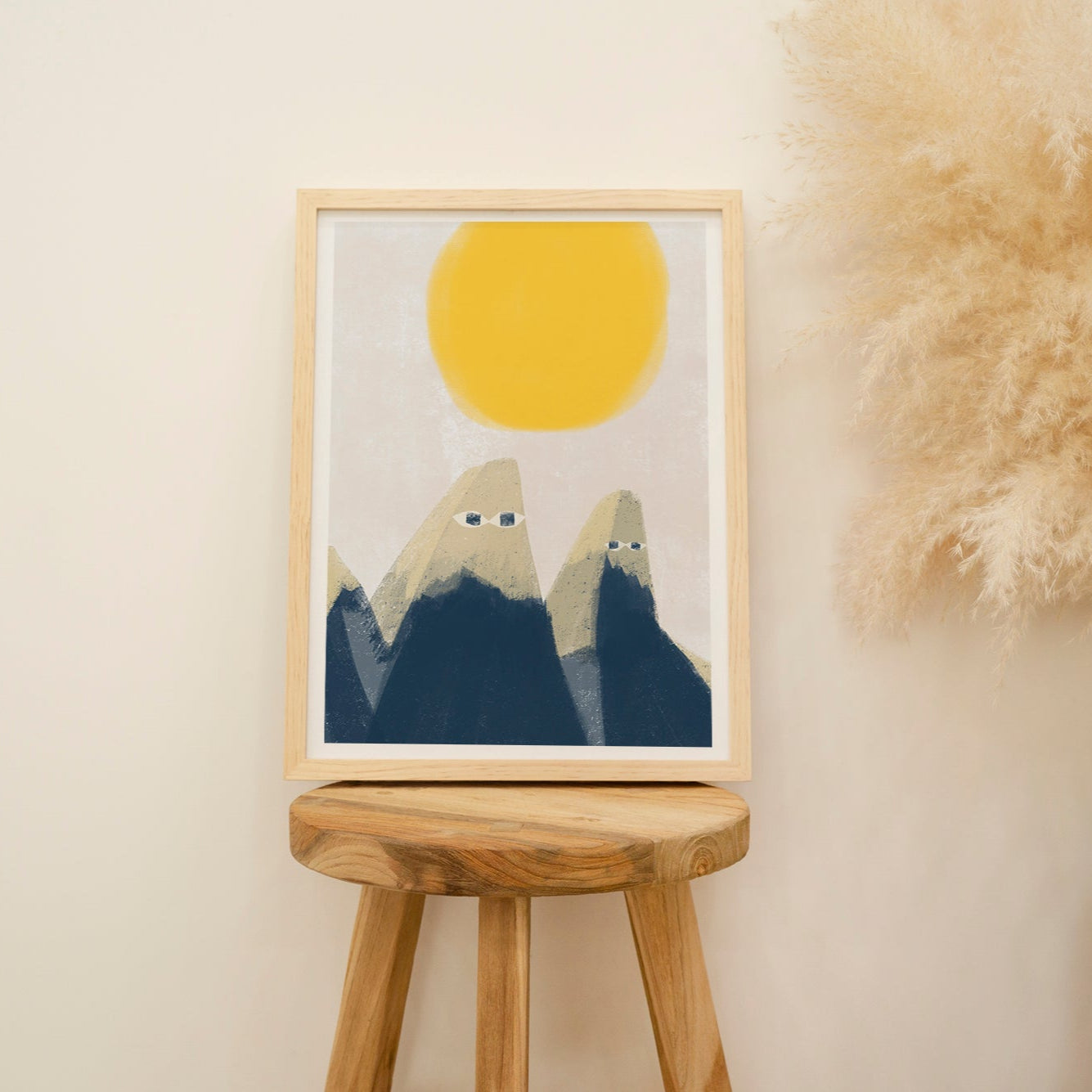 Framed artwork with mountains and a yellow sun on a wooden stool against a beige wall.