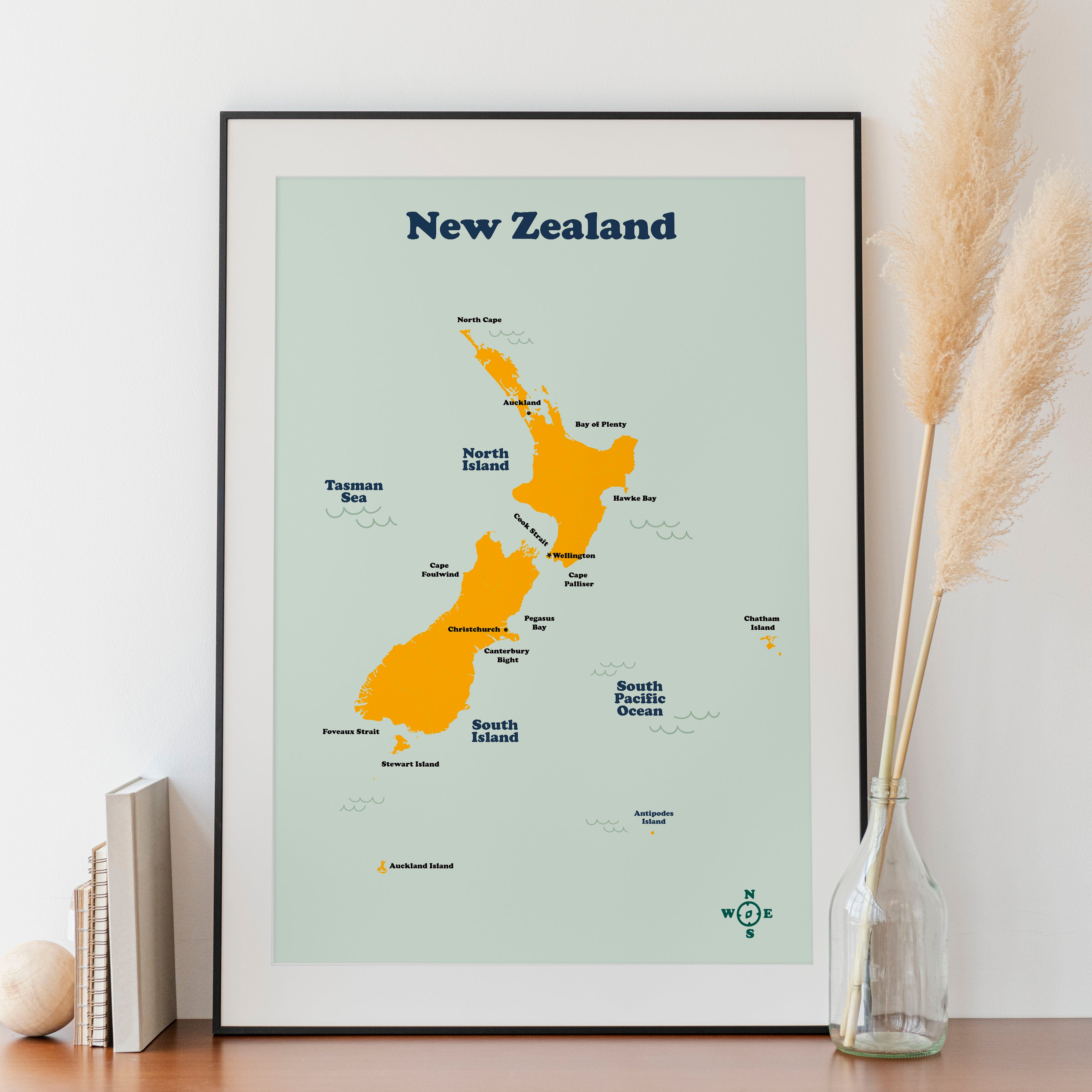 A framed map of New Zealand displayed on a white surface with a decorative dry plant beside it.