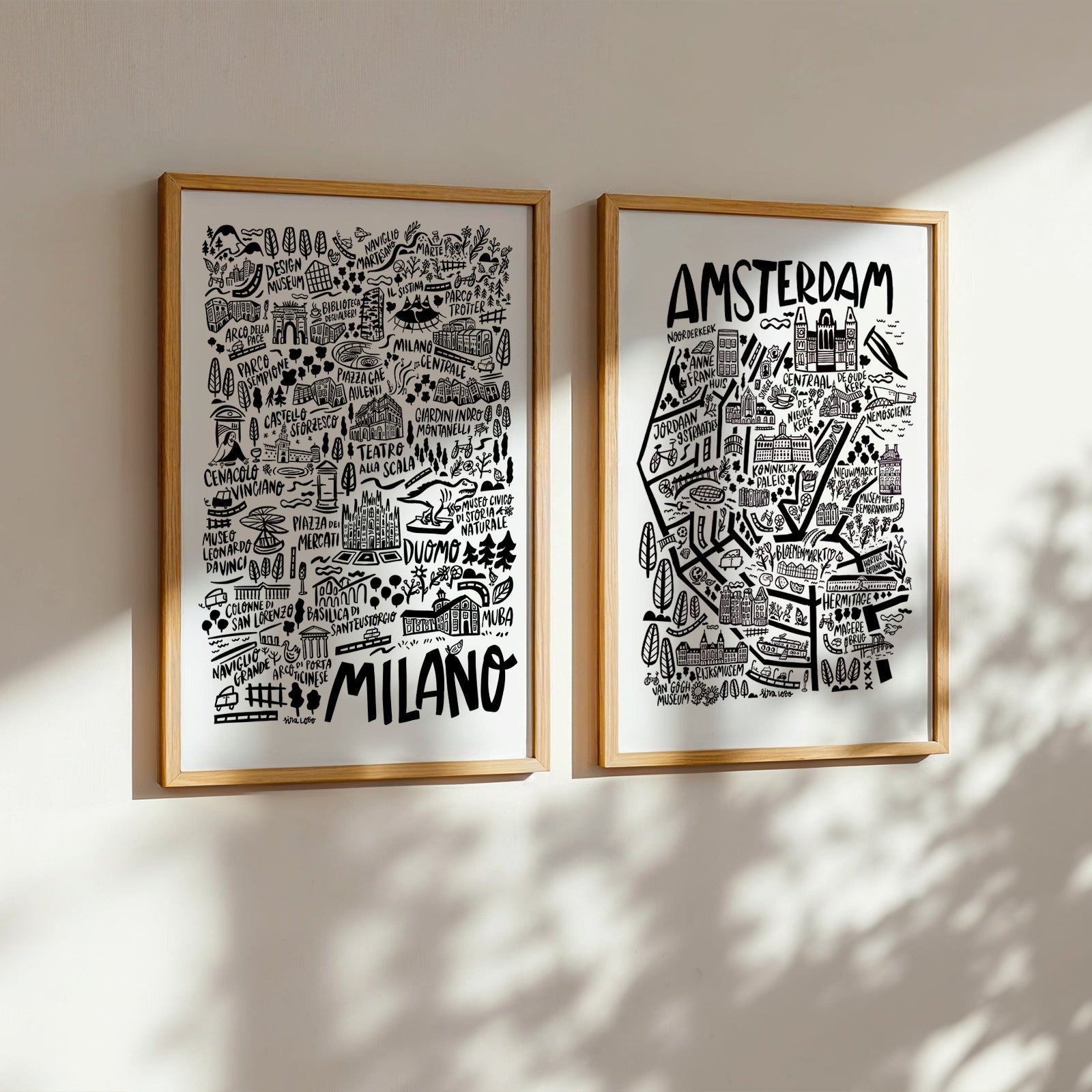 Two framed maps of Milano and Amsterdam on a wall with sunlight casting shadows.