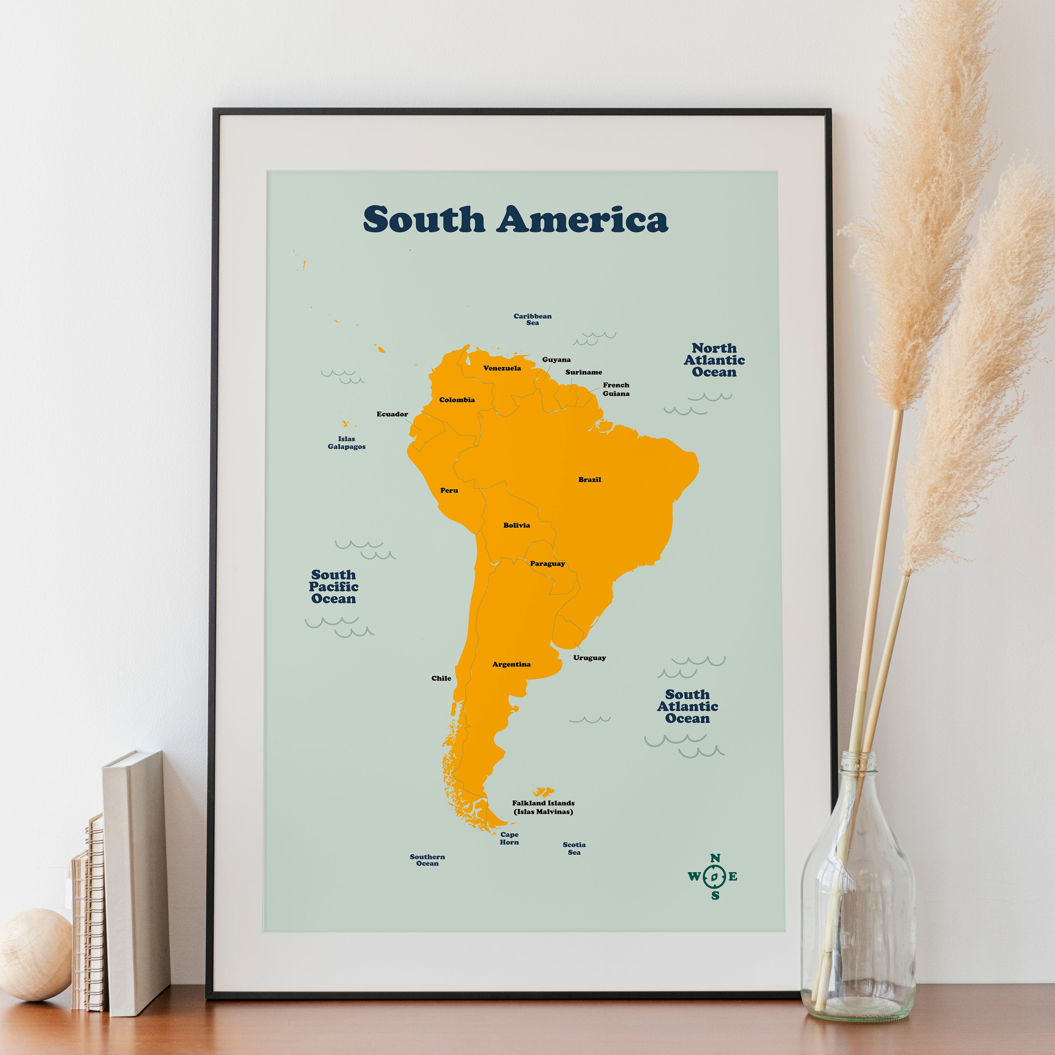 A framed map of South America displayed on a white surface with a decorative vase and a book in the background.