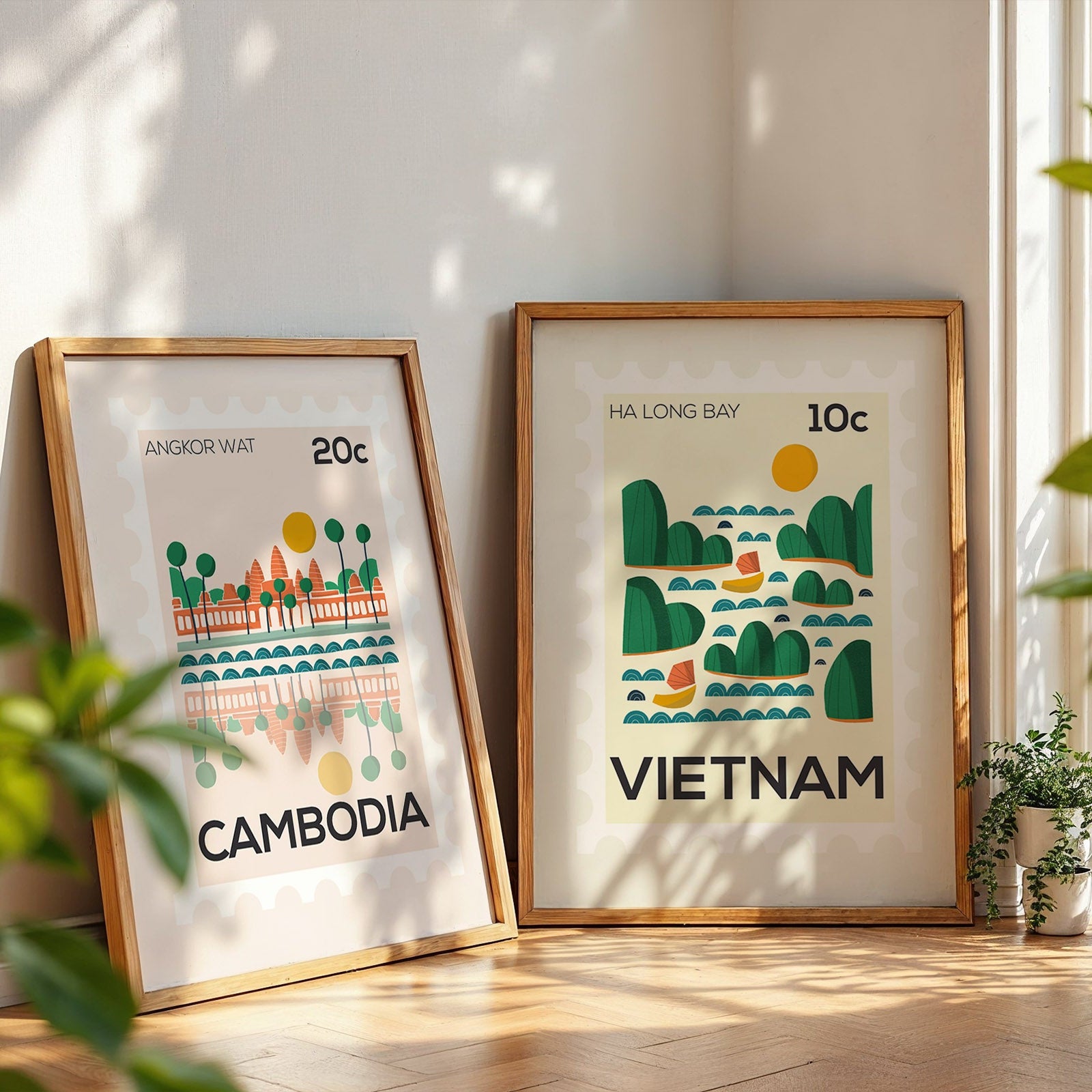 Two framed prints of Cambodia and Vietnam on a wooden floor with plants around.