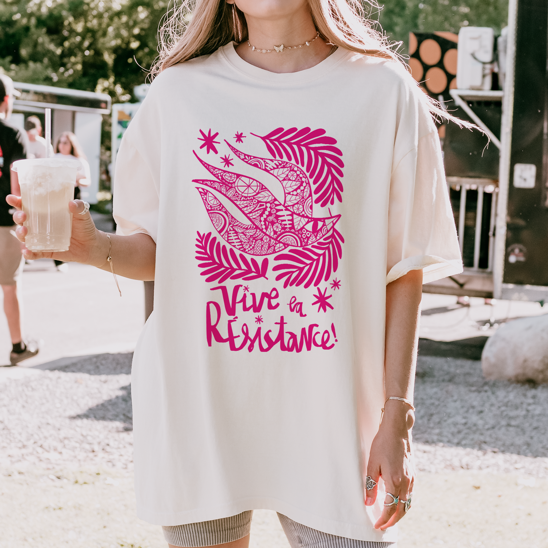 Person wearing a white t-shirt with pink graphic and text, holding a drink outdoors.