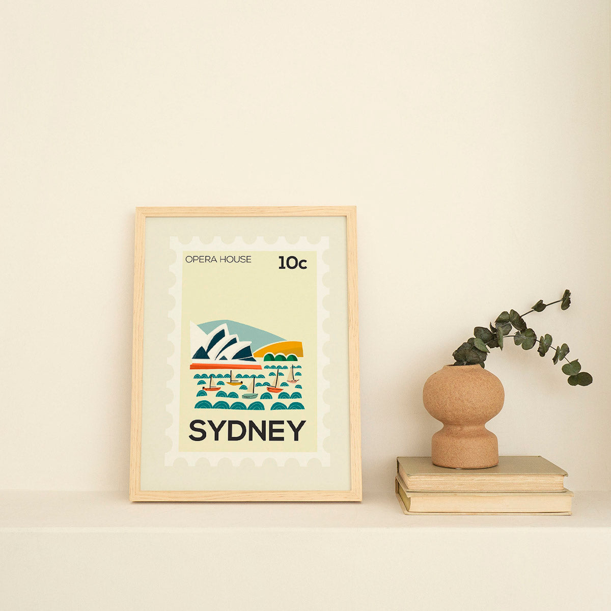 Framed artwork of the Sydney Opera House with a vase and books on a beige surface.