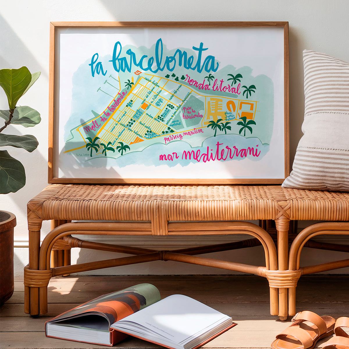 Framed map of La Barceloneta on a wicker table with books and sandals.