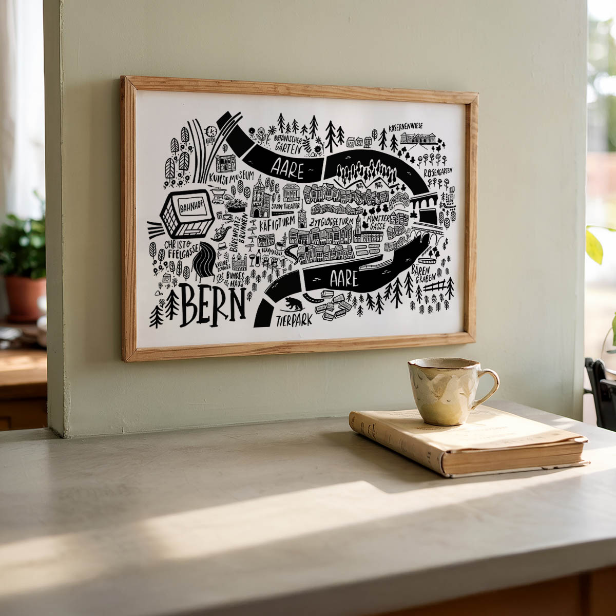 Framed black and white map of Bern on a wall above a table with a book and mug.