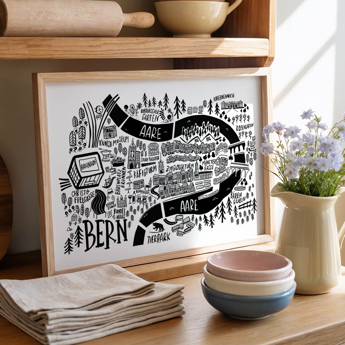 Framed map print on a kitchen counter with bowls, flowers, and a towel.