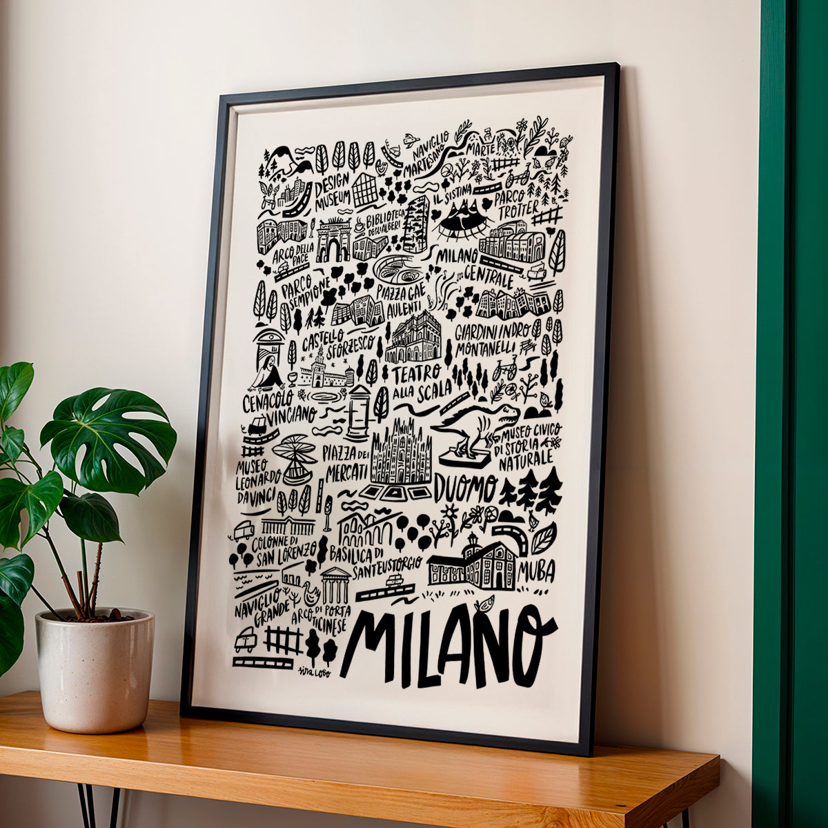 Framed artwork with 'Milano' on a wall next to a plant