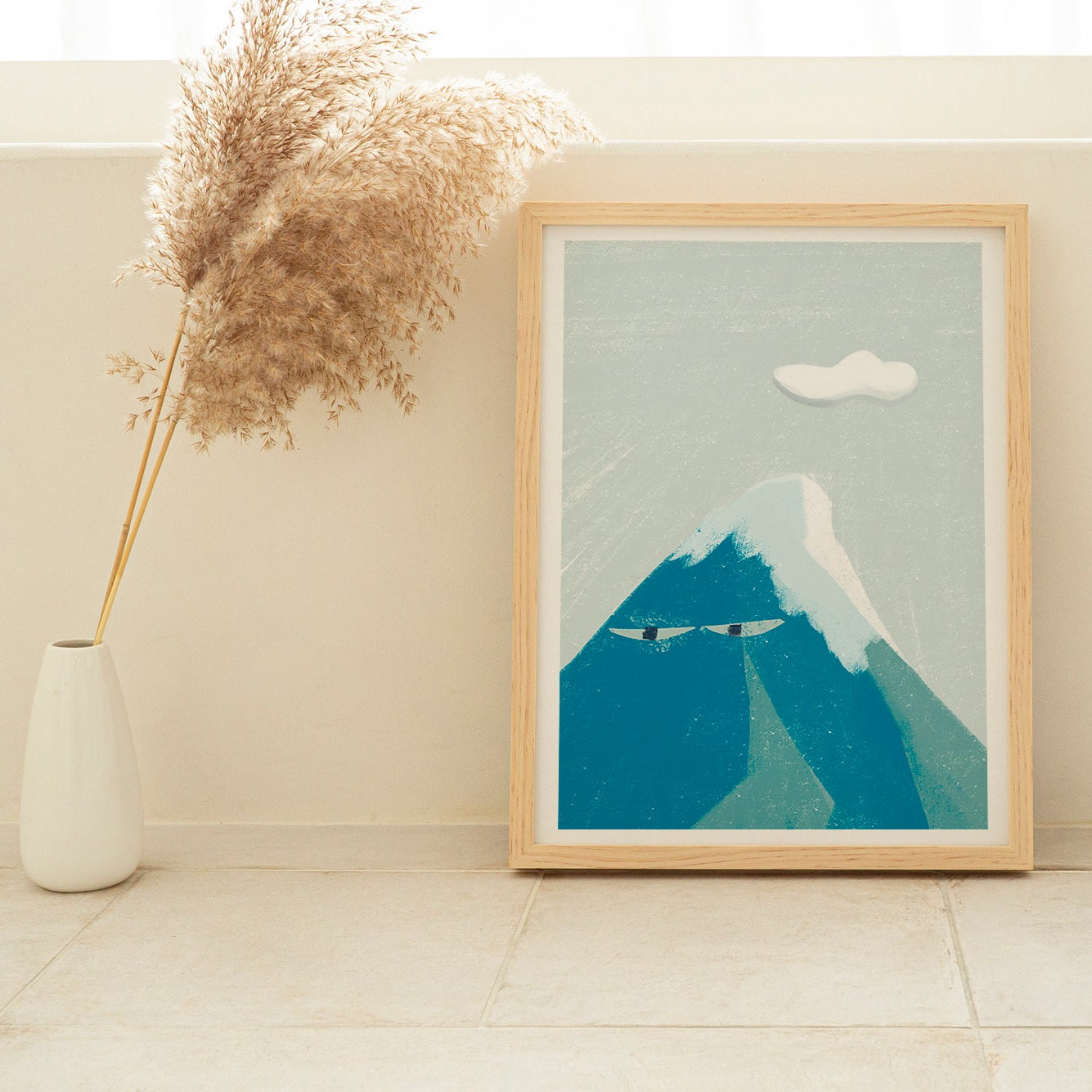 Framed artwork of a mountain on a white wall with a vase and dried plants.