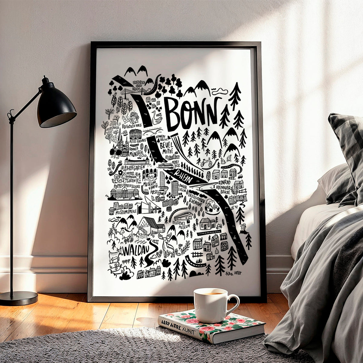 Framed black and white map of Bonn on a wall in a room with a lamp and book.