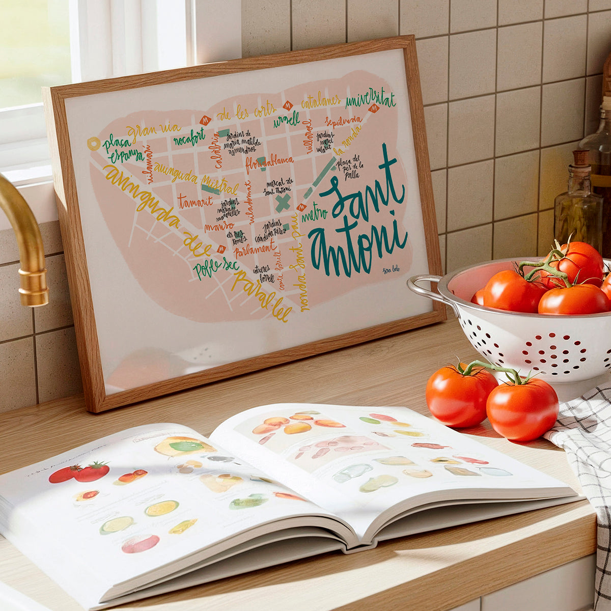 Framed map of Sant Antoni on a kitchen counter with an open book and tomatoes.