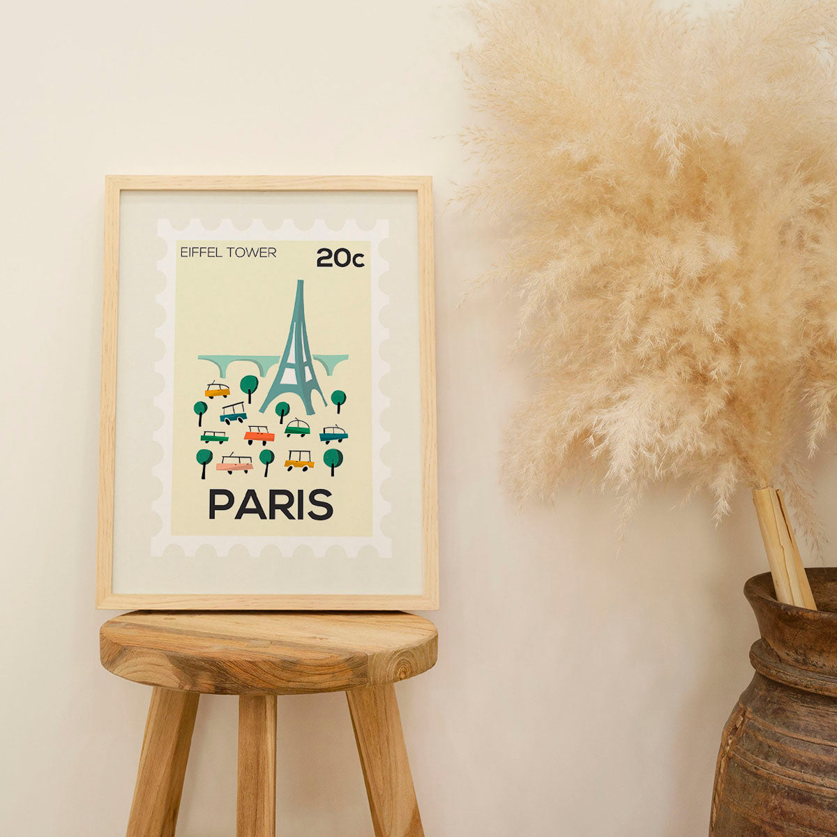 Framed artwork of the Eiffel Tower with 'PARIS' text on a wooden stool against a beige wall.