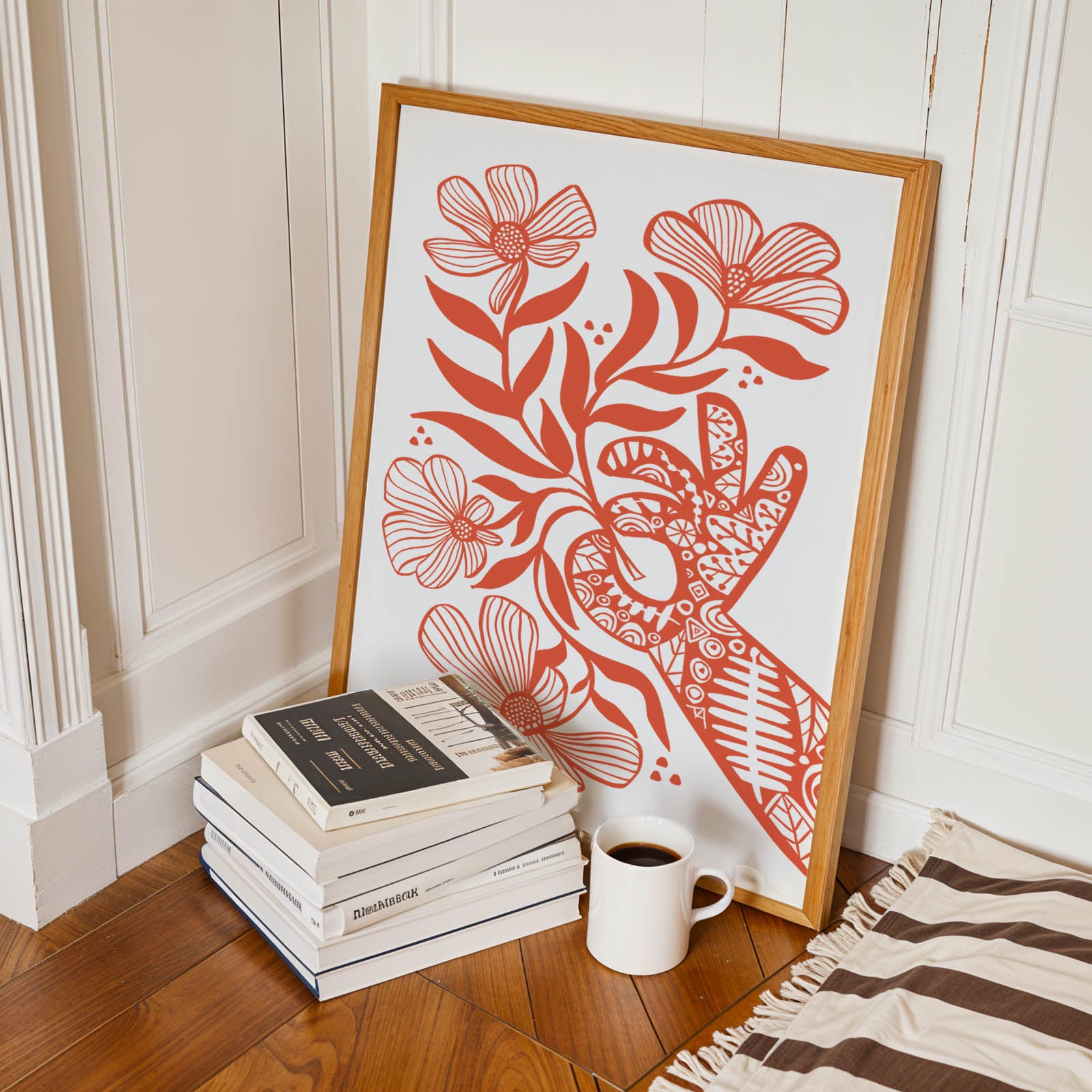 Framed floral artwork on a stack of books with a cup next to it on a wooden floor.