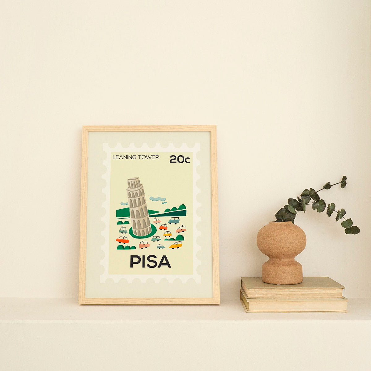 Framed artwork of the Leaning Tower of Pisa on a beige wall with a vase and books.