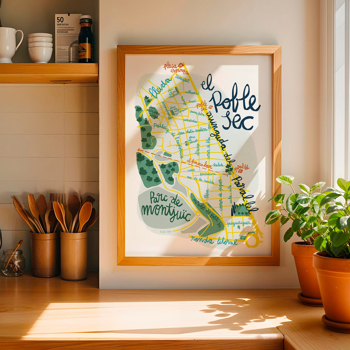Framed print of poble Sec in Barcelona on a kitchen wall with plants and utensils below