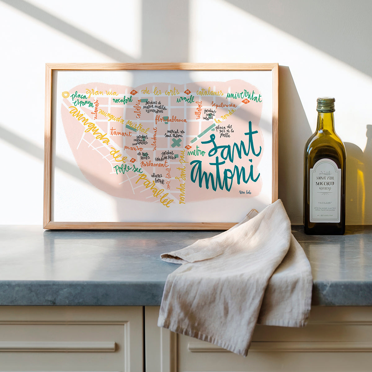 Framed artwork with colorful text on a kitchen counter next to a bottle of olive oil.