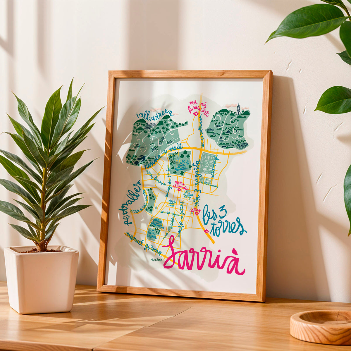 Framed map of Sarrià in Barcelona with colorful text on a wooden table, surrounded by plants.