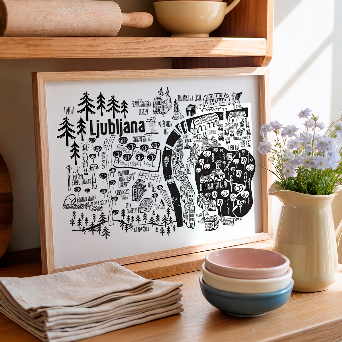 Framed map of Ljubljana on a wooden shelf with kitchen items and flowers.