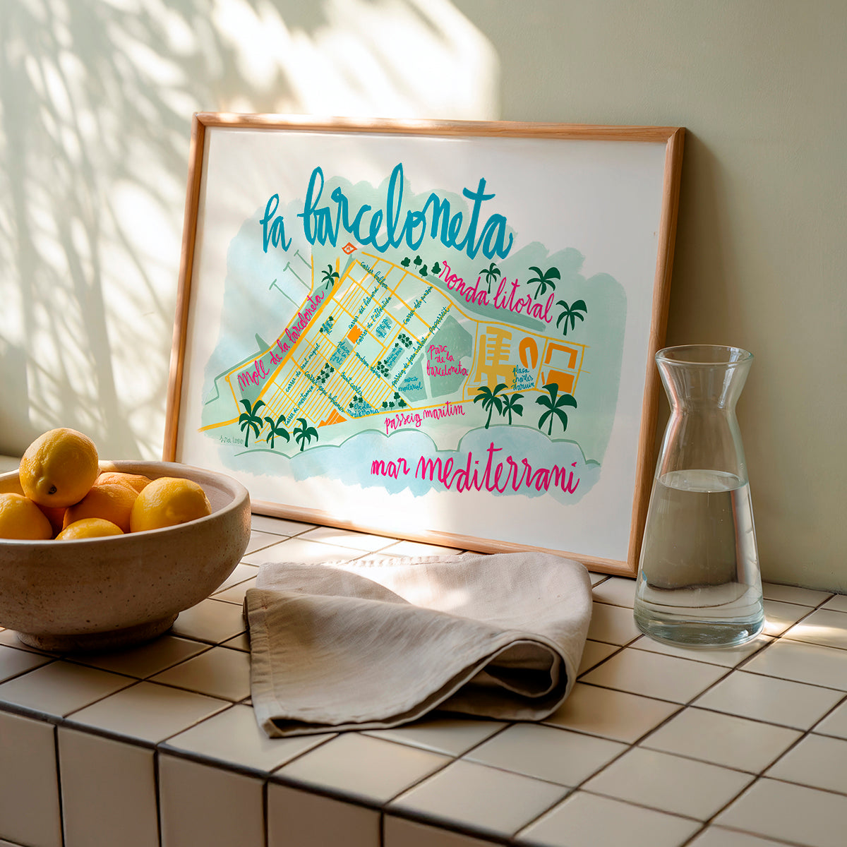 Framed map of La Barceloneta with a bowl of lemons and a carafe on a tiled surface.