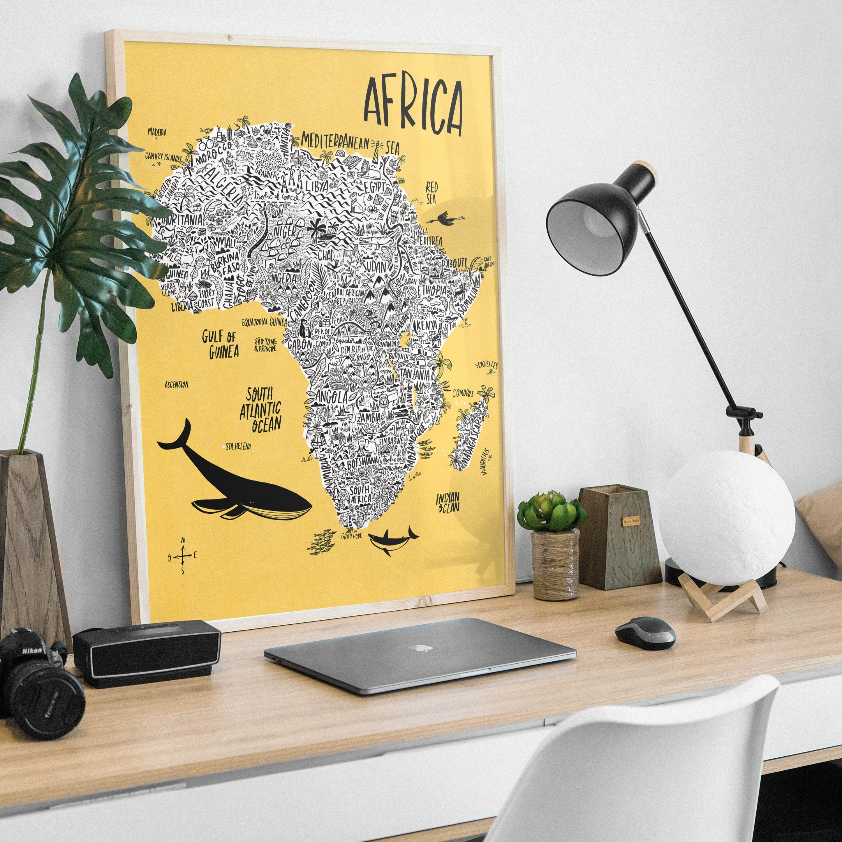 African continent map poster with a yellow background, featuring various illustrations such as a whale, a giraffe, and a map outline, displayed on a desk with a laptop, a camera, and a plant in the background.