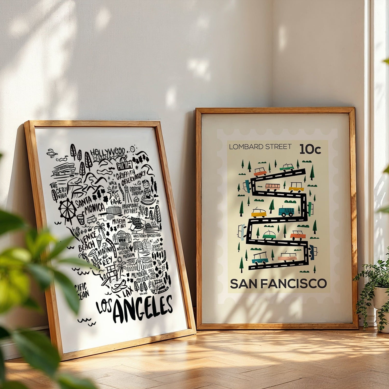 Two framed maps of Los Angeles and San Francisco on a wooden floor with plants around.