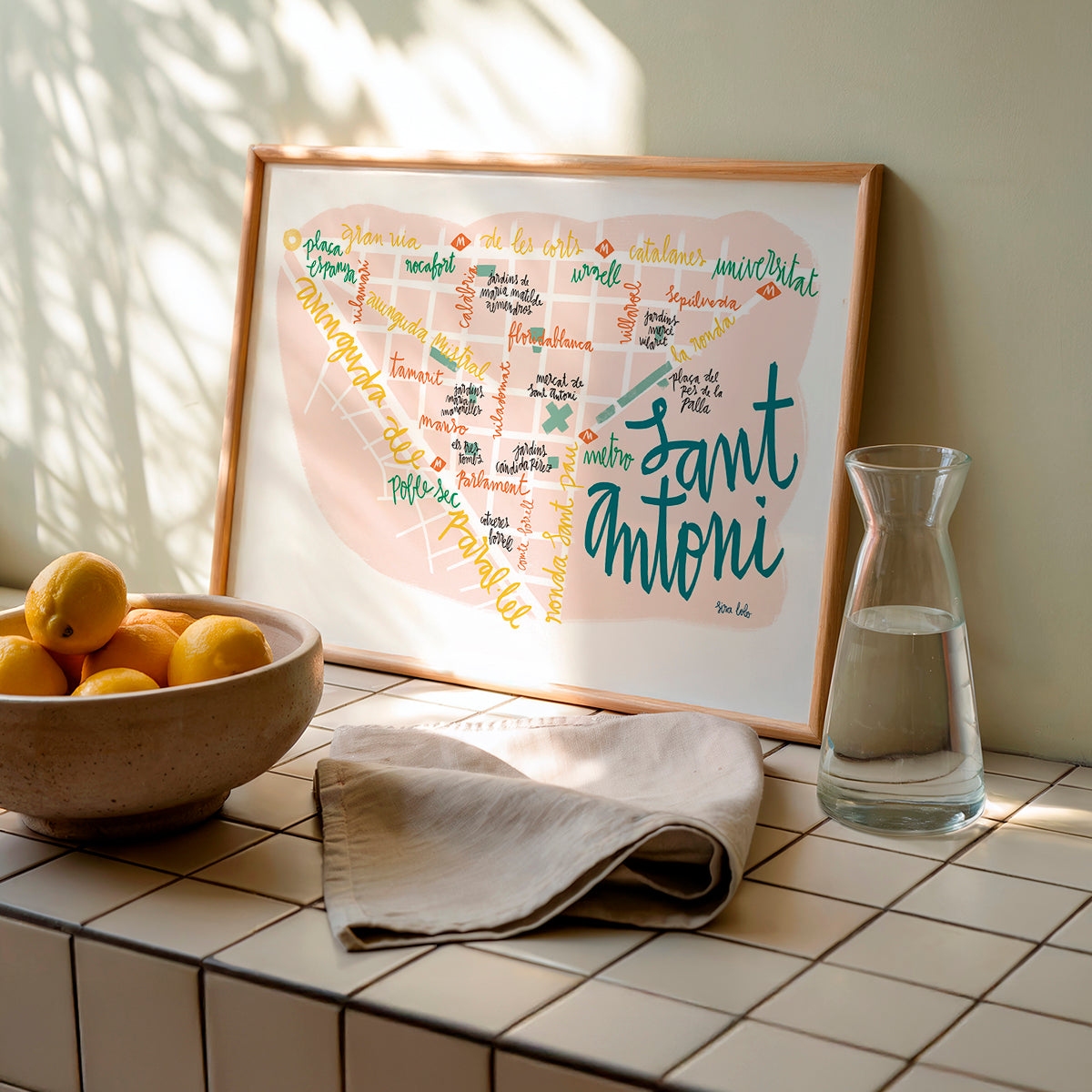 Framed map of Sant Antoni on a tiled surface with a bowl of fruit and a glass carafe.