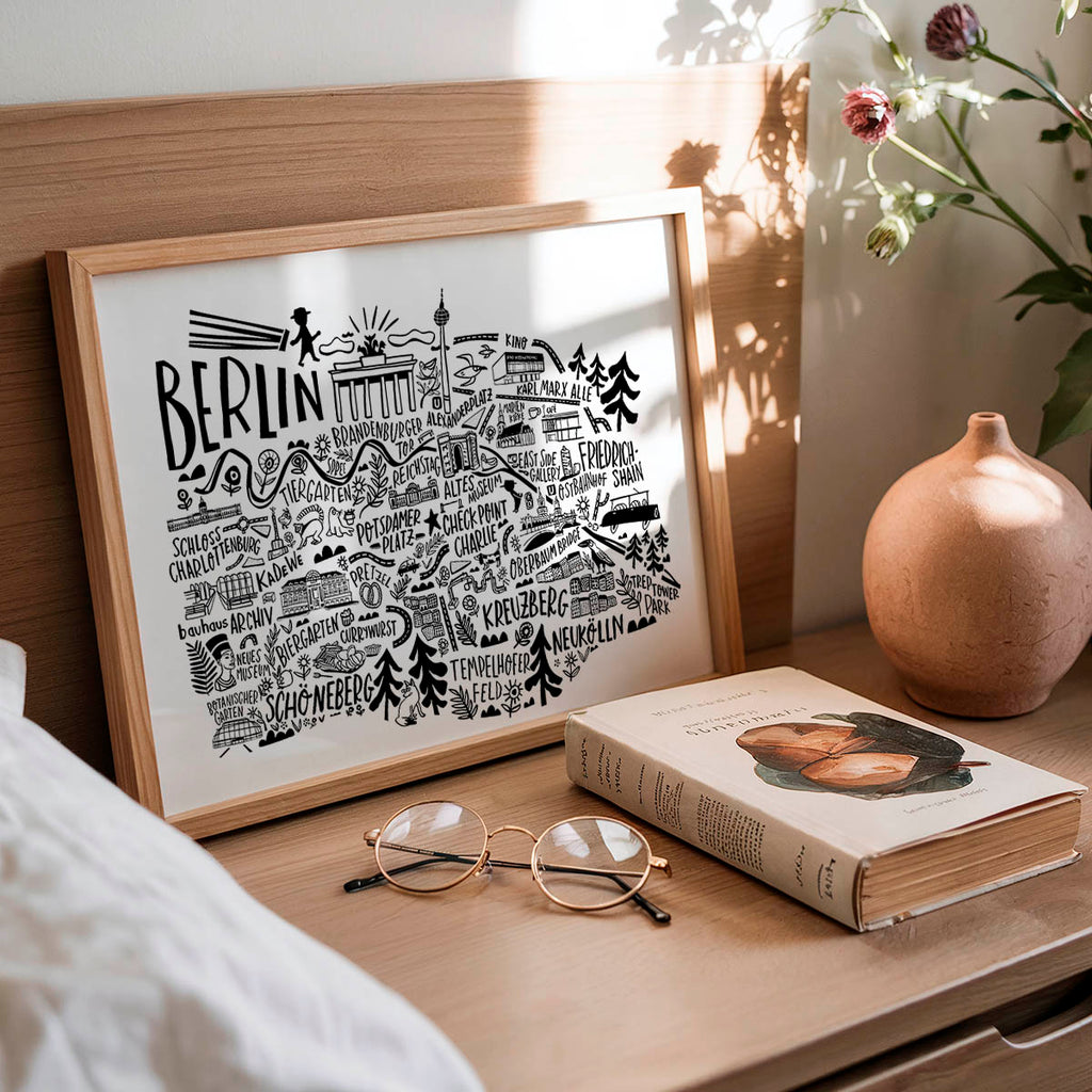 Framed artwork with 'Berlin' design, book, and glasses on a wooden surface.