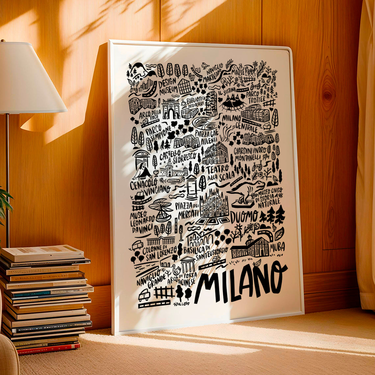 Framed map of Milano with black illustrations on a white background, leaning against a wooden wall.