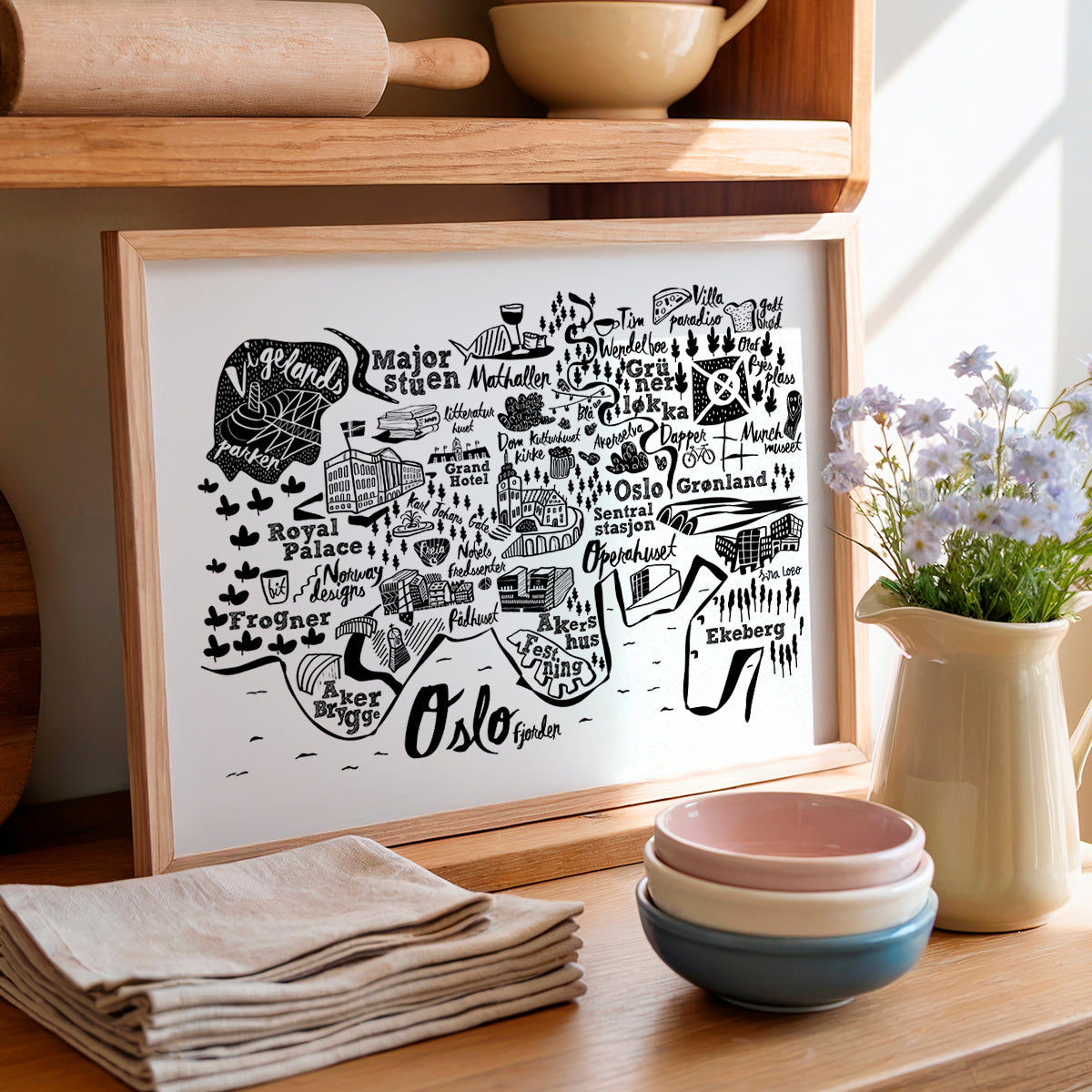 Framed map of Oslo on a kitchen shelf with bowls, flowers, and a towel.